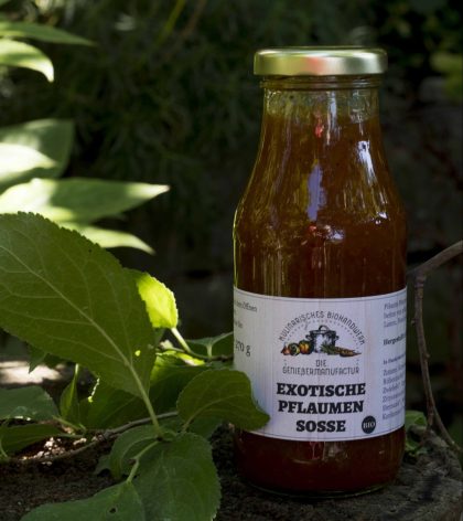 Exotische Pflaumensoße; eine Flasche mit goldenem Deckel beinhaltet die Soße. Die Flasche ist mit einem weißen Etikett versehen, auf dem Logo und Beschriftung ist. Im Hinergrund sind grüne Blätter zu erkennen.