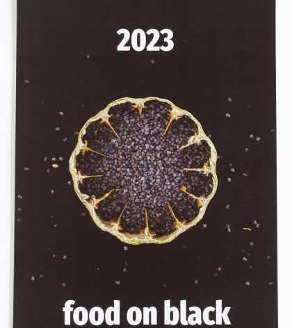 food on black - Kalender 2023; Das Titelblatt zeigt eine aufgeschnittene Mohnkapsel mit Mohn darin auf schwarzem Hintergrund. Einige Mohnsamen liegen dekorativ verstreut um die Mohnkapsel herum. Oben steht die Jahreszahl 2023, unten der Begriff food on black Kalender