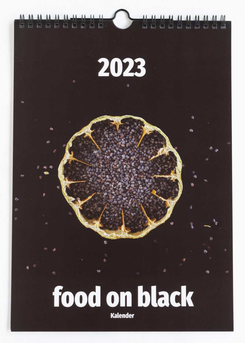 food on black - Kalender 2023; Das Titelblatt zeigt eine aufgeschnittene Mohnkapsel mit Mohn darin auf schwarzem Hintergrund. Einige Mohnsamen liegen dekorativ verstreut um die Mohnkapsel herum. Oben steht die Jahreszahl 2023, unten der Begriff food on black Kalender
